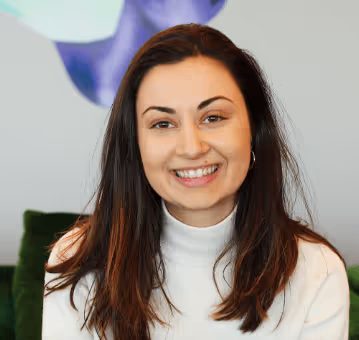 Woman with long dark hair in a white turtleneck, smiling, against a soft geometric background