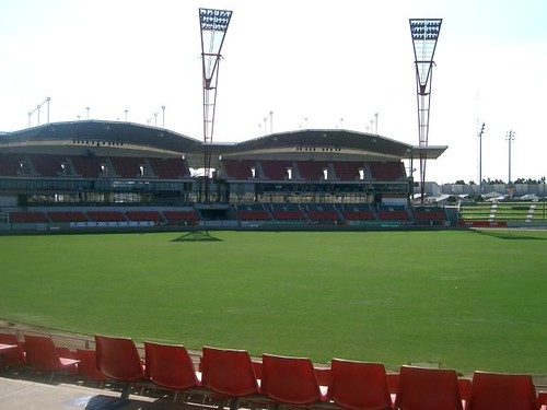Empty green sports field with red seats and two floodlight towers under a bright sky